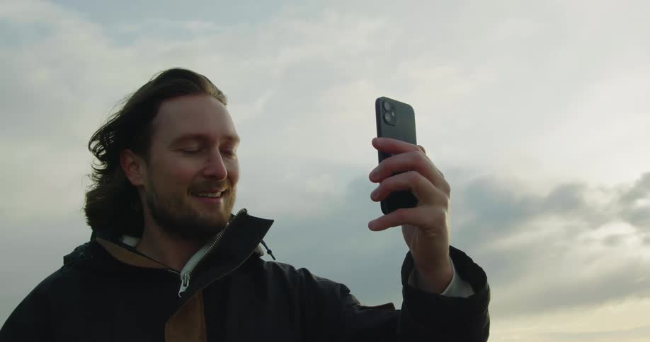 A man standing on a beach calls someone on his cell phone and waves as he video chats with them