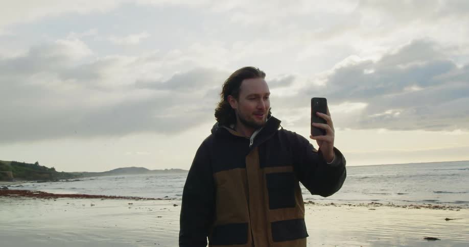 A man waves and smiles as he talks on a video call standing on the beach