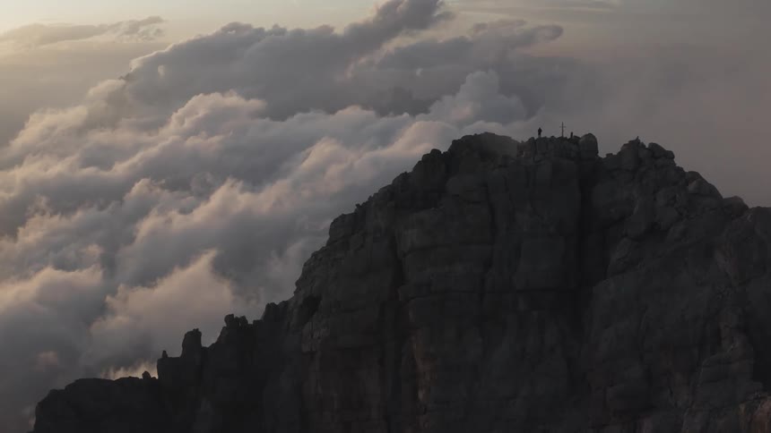 Person stands next to a cross on top of a mountain under the clouds