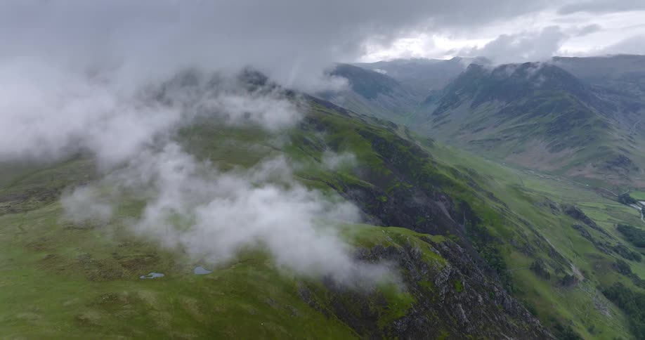Clouds hang low over a grass covered mountain ridge and there are mountains in the distance