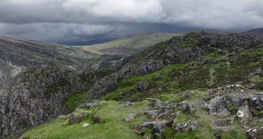Grass grows on a rocky mountain ridge and there are mountains on the horizon under a dark cloudy sky