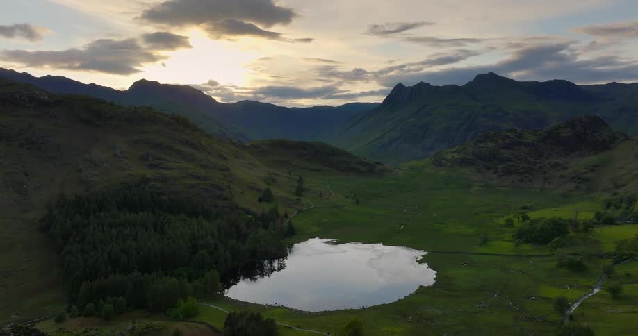 Sun shines from over mountains onto valley containing woods a winding river and a reflective pond
