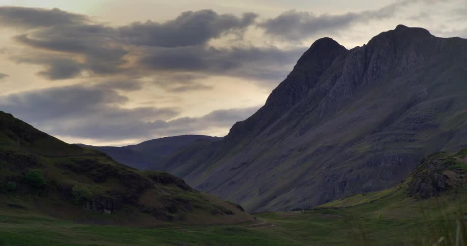 Beyond a grass covered slope is a mountain range with peaks high in a cloudy sky