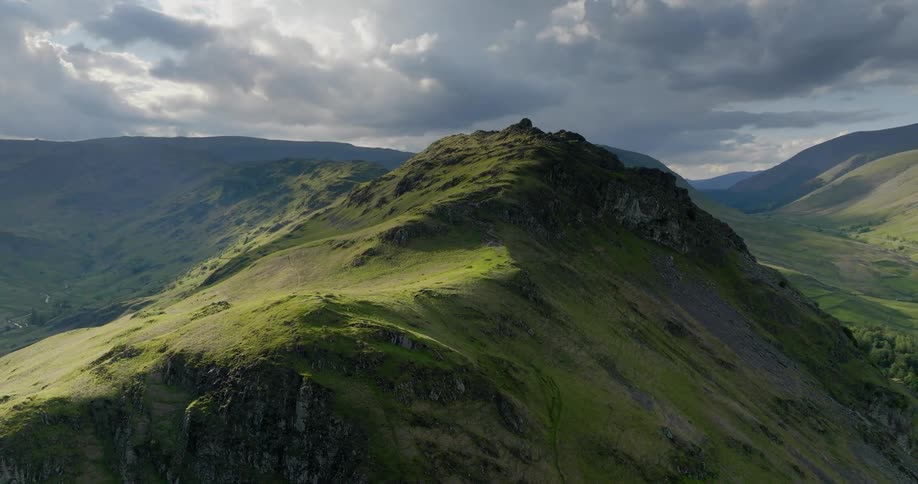 The sun shines on a grass covered mountain ridge and there are mountains in the distance under a cloudy sky