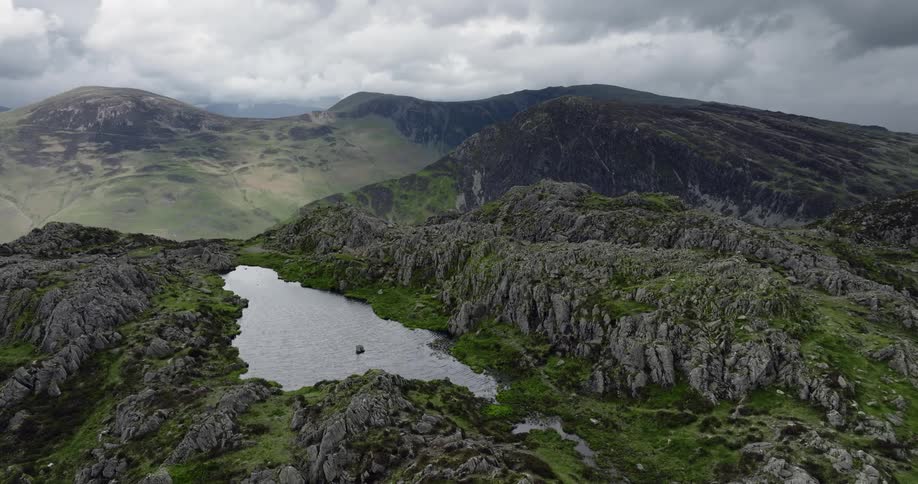 Grass grows on a rocky mountain ridge with a lake and there are mountains in the distance under a cloudy sky