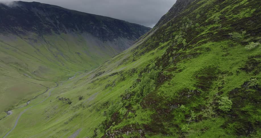 River and road run through narrow valley formed by green covered mountain slopes under an overcast sky