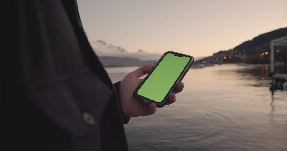 A man is standing near a lake holding a cell phone with a green screen and there is a snow covered mountain in the distance