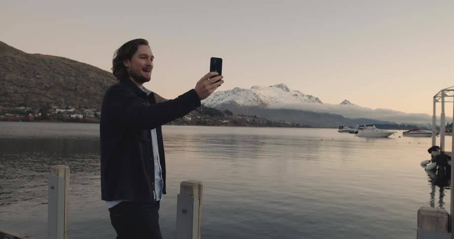 A man stands on the shore and holds a cell phone and waves as he makes a video call