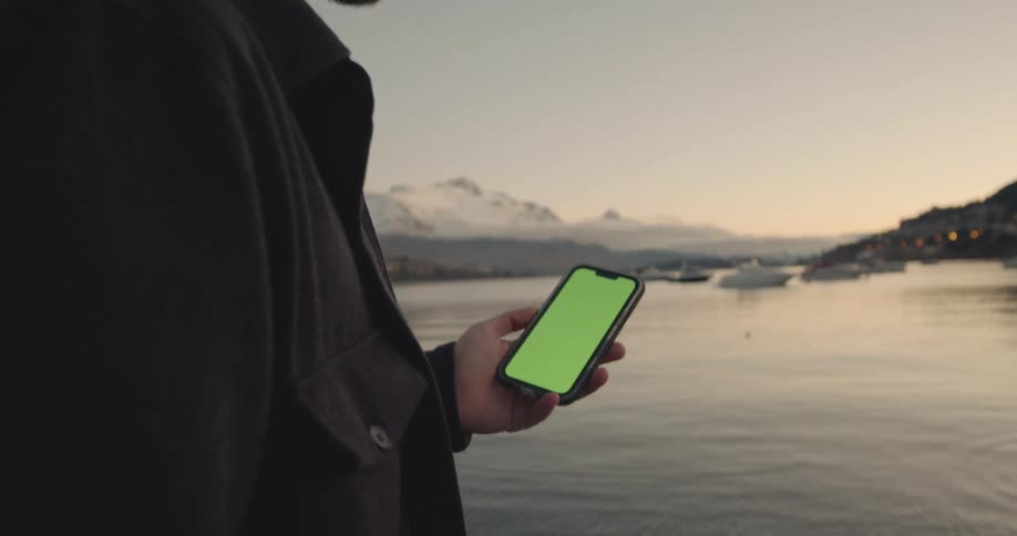 A man looks at his cell phone as he stands next to a lake in the mountains that has several boats in it