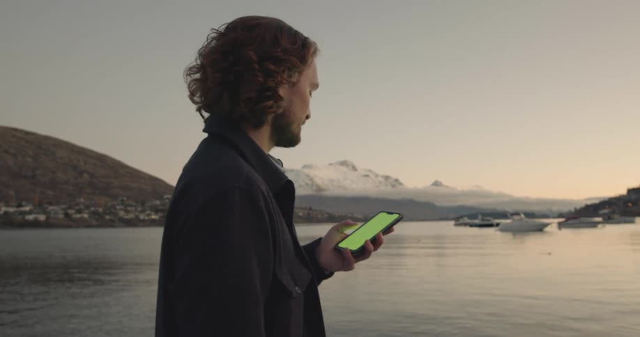 A man taps the screen of his smartphone with his thumb while standing on the coast during the day