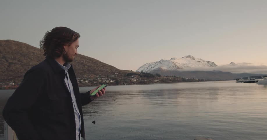 A man looking out over a body of water while standing on the shore checks his phone before returning his gaze to the water