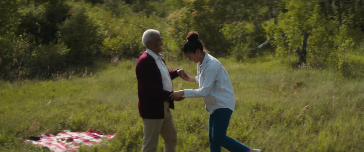 A young woman is dancing in a field near the woods with an elderly man