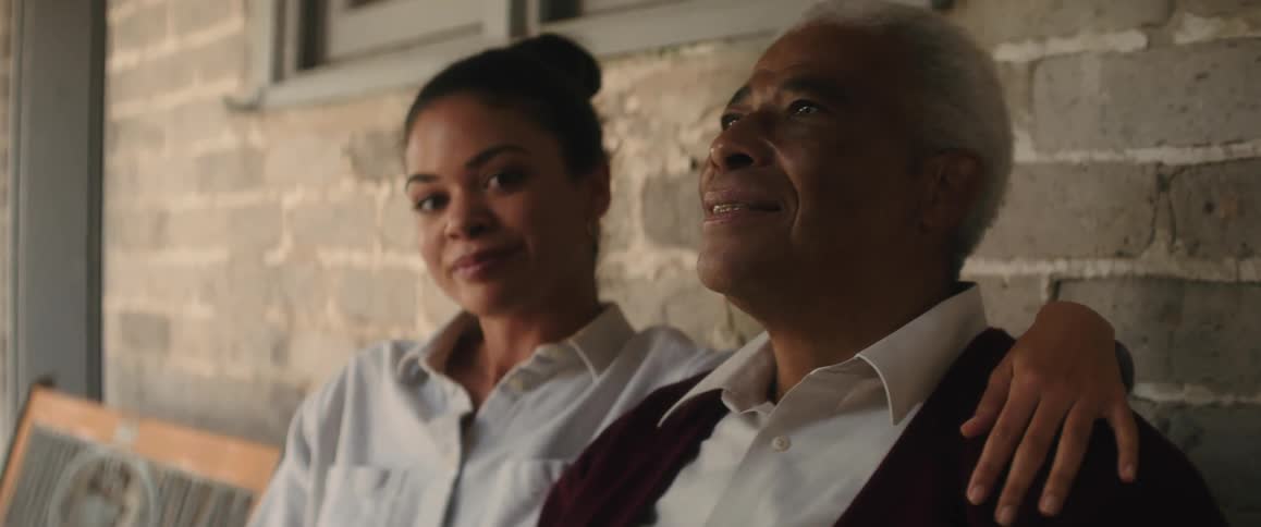A young woman is sitting on a bench outside of a building with her arm around an elderly mans shoulders as she smiles at him
