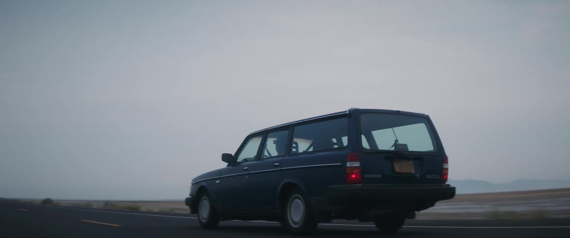 An old blue volvo station wagon is traveling on a road along the coast and there are mountains in the distance