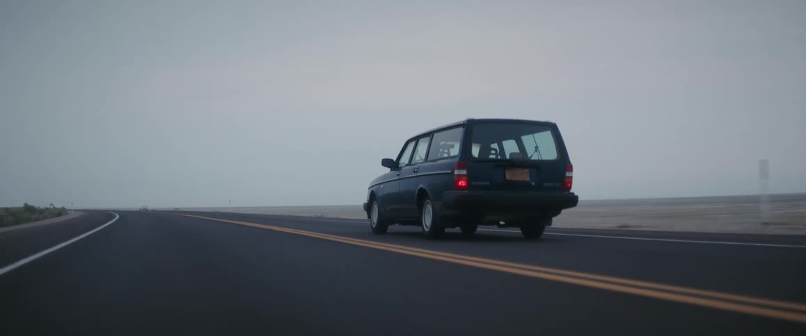 A person drives a volvo station wagon along a road through a flatland on a cloudy day