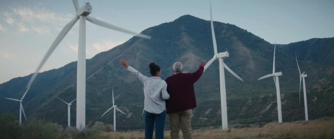An elderly man and a young woman are standing outside at a wind farm raising their hands in the air as the wind turbines spin and there is a mountain in the distance