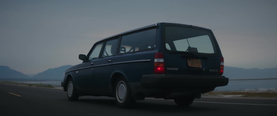 An old blue volvo station wagon is traveling on a road along the coast and there are mountains in the distance