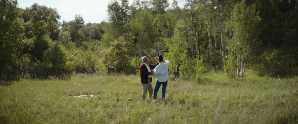 A young woman is dancing in a field near the woods with an elderly man