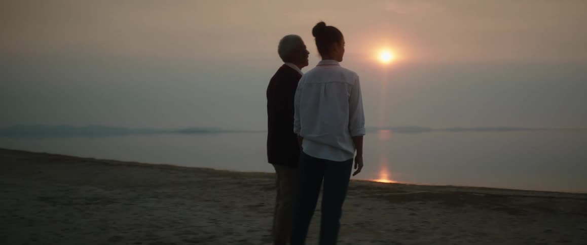 Woman with bun and white shirt stands next to man with black blazer shirt and pants on the coast at sunset