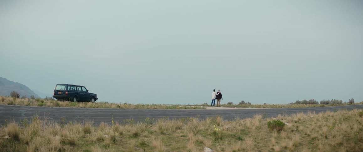 A woman walks away from a car on the side of the road with her arm around her elderly father and points in to the distance
