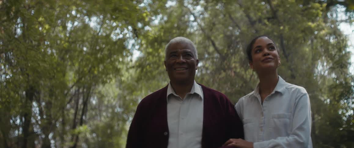 A woman holds her fathers arm as they walk under tall trees and talk