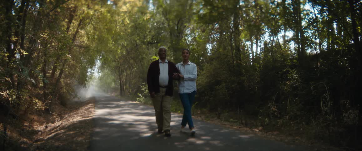 A young woman is holding the arm of an elderly man as they walk on a road through the woods
