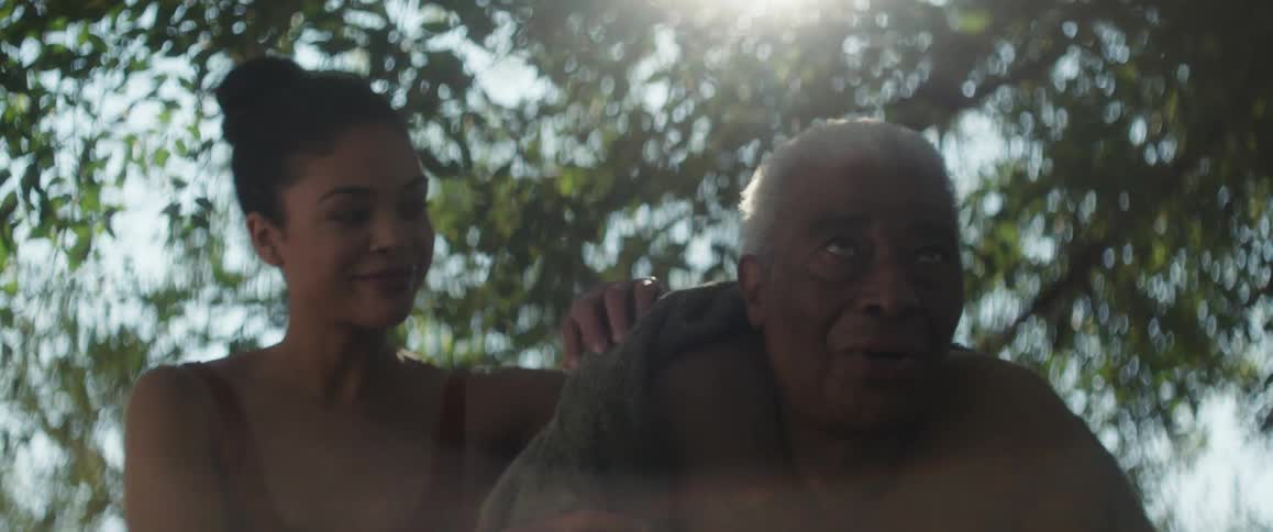 A woman in a swimsuit puts a towel around the shoulders of an elderly man sitting next to her outdoors during the day