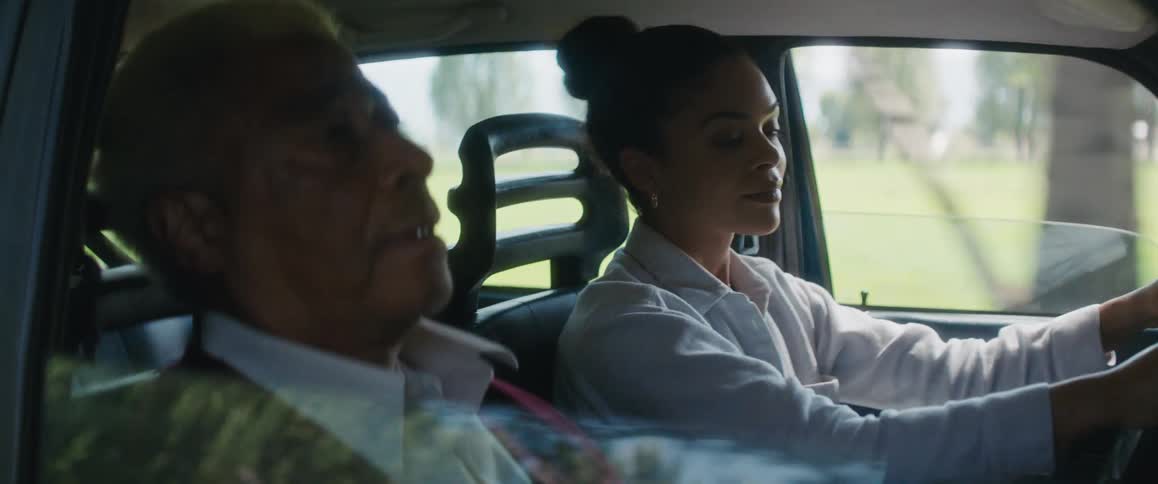 A woman driving a car in a rural area looks over at her father in the passenger seat and smiles