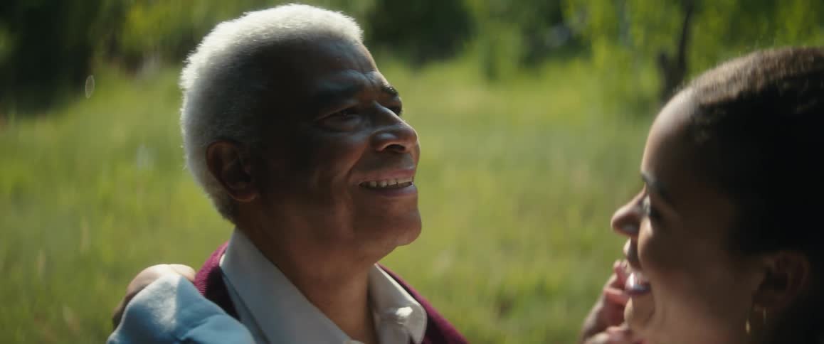 An elderly man and a young woman are standing outside near the woods dancing and smiling as they talk to each other