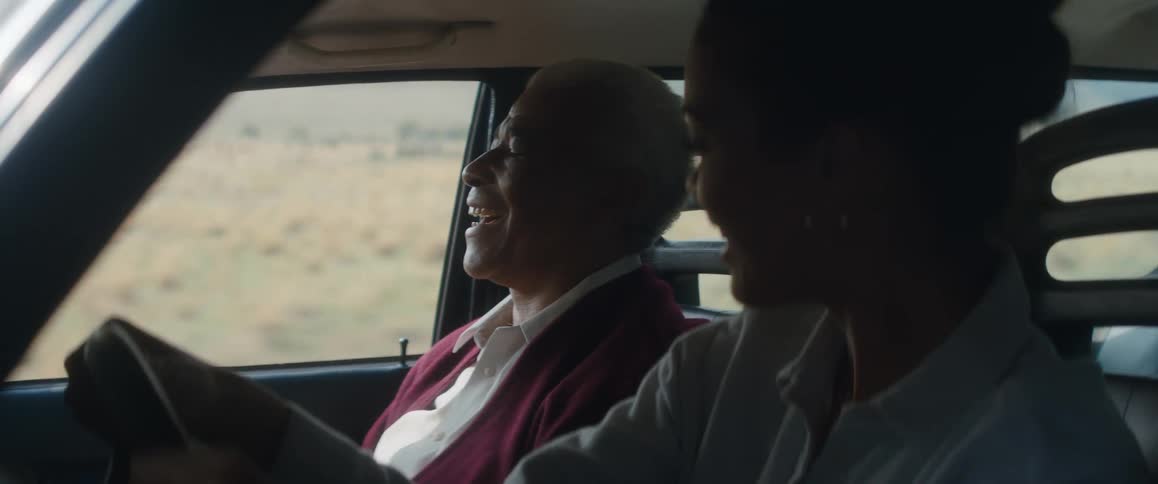 A young woman is driving an automobile and she is singing a song with the elderly man in the seat next to her