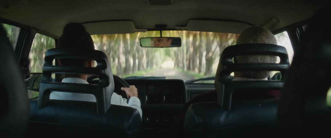 A woman steering the car on a tree lined driveway turns to speak to her grandfather