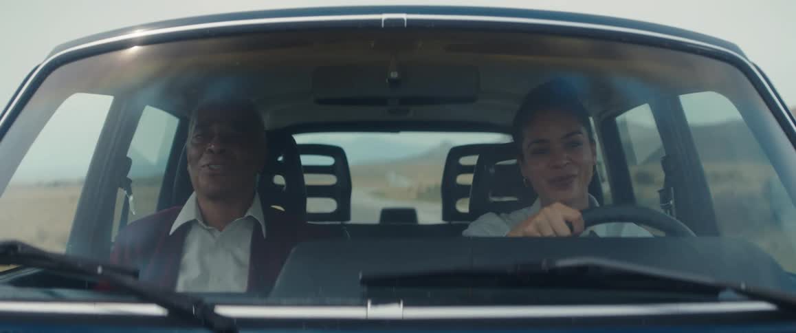 A woman and an elderly man sing together while traveling in a vehicle before bursting into laughter