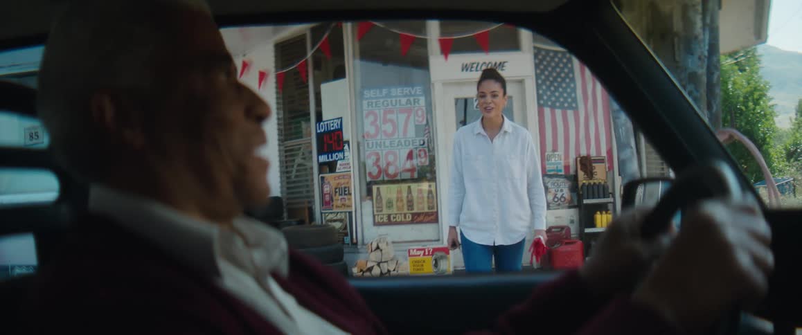 A old man sits in a car as his daughter walks away from a country store and looks up surprised and happy to see him and goes to him