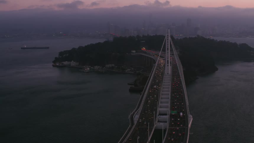 Traffic flows over suspension bridge to and from city skyline under a red sky