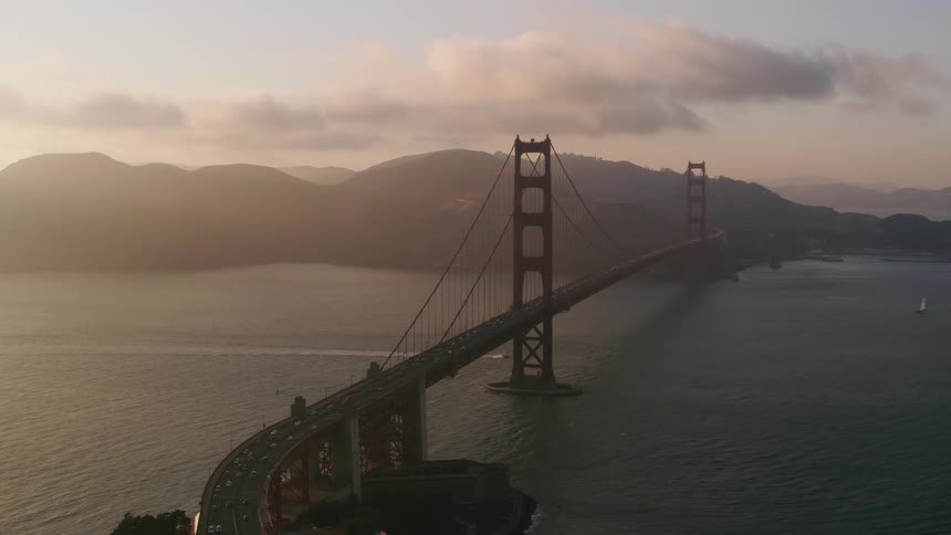 Traffic moves across a suspension bridge as a boat sails below and there are mountains in the distance under a cloudy and foggy sky