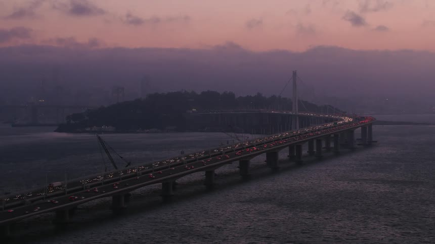 Traffic moves across a bridge stretching across the water from the coast with headlights on under a dark cloudy red sky