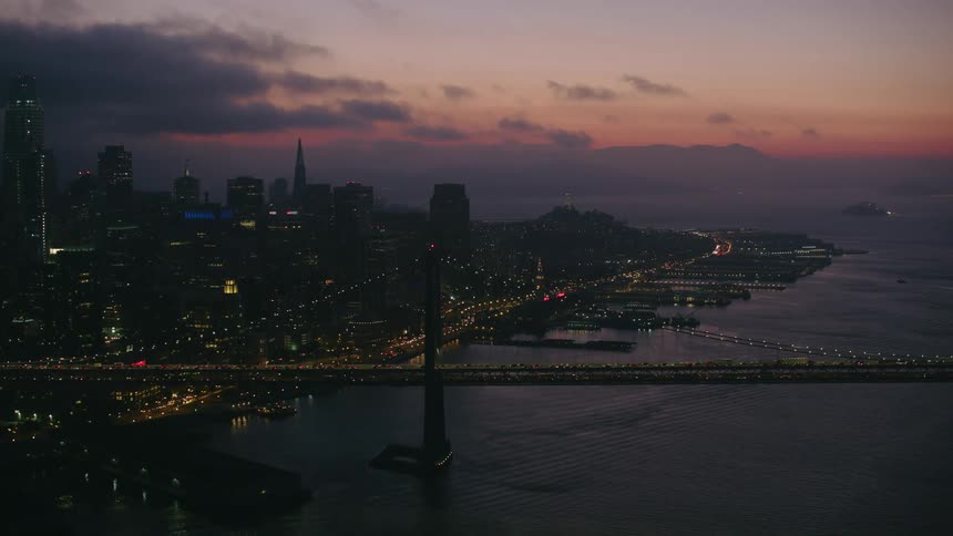 A big city along the coast with lights lit up as traffic moves across a suspension bridge and tall buildings and skyscrapers reach high into a dark colorful sky