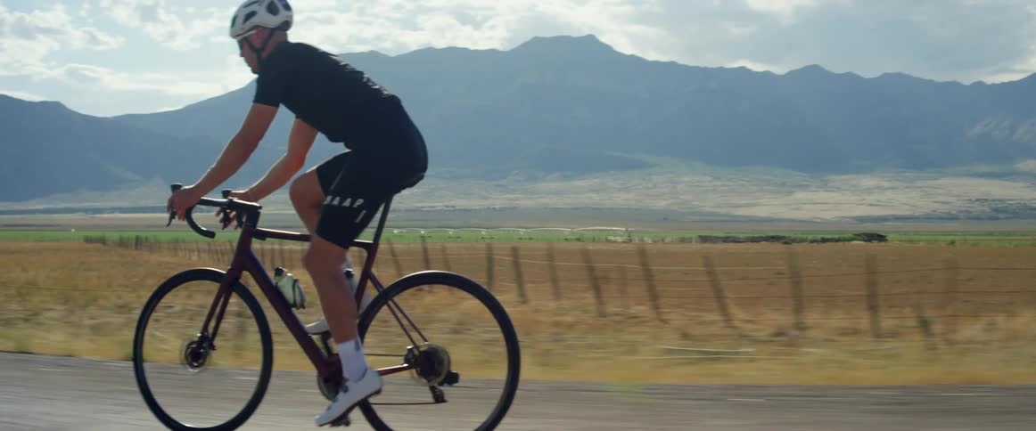 A man wearing a helmet is riding a bicycle on a rural road with mountains in the distance