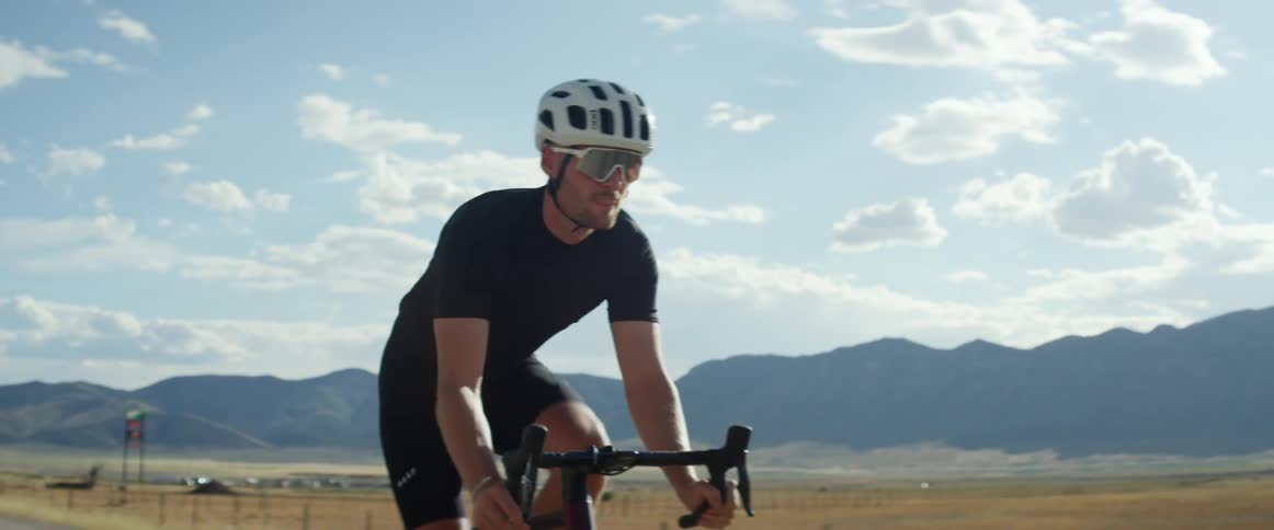 Male cyclist pedals along road past fenced pasture with mountains in the background