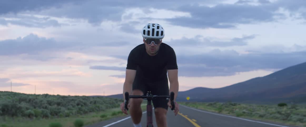 Man rides a bicycle on a highway as cars approach behind him and mountains sit in the distance