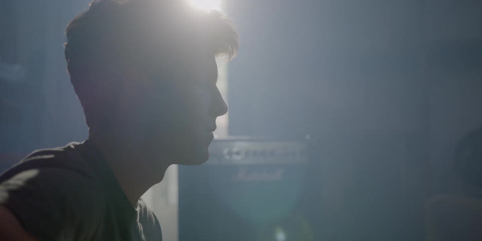 A teenage boy sits in his room and nods his head to music as he plays a guitar