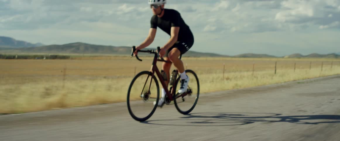 Man wearing white shoes a helmet and sunglasses rides a bicycle on a road next to a field on a sunny day