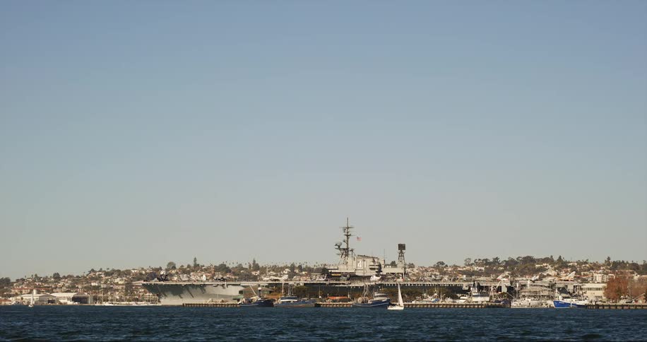 Sailboat traveling in the lake with ships docked nearby under a blue sky