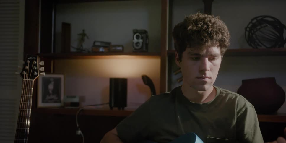 A man plays the guitar while sitting indoors