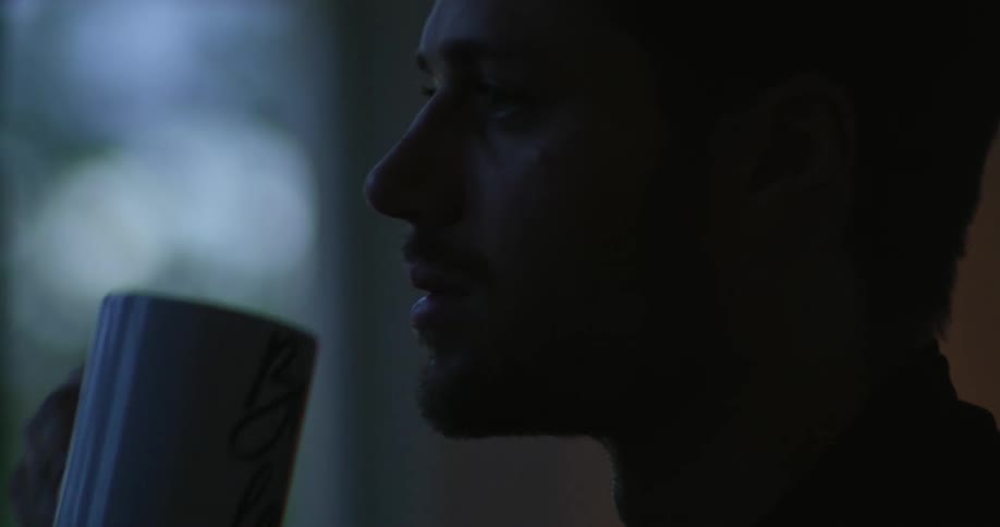 A man scratches his chin while drinking coffee in a dimly lit room during the day