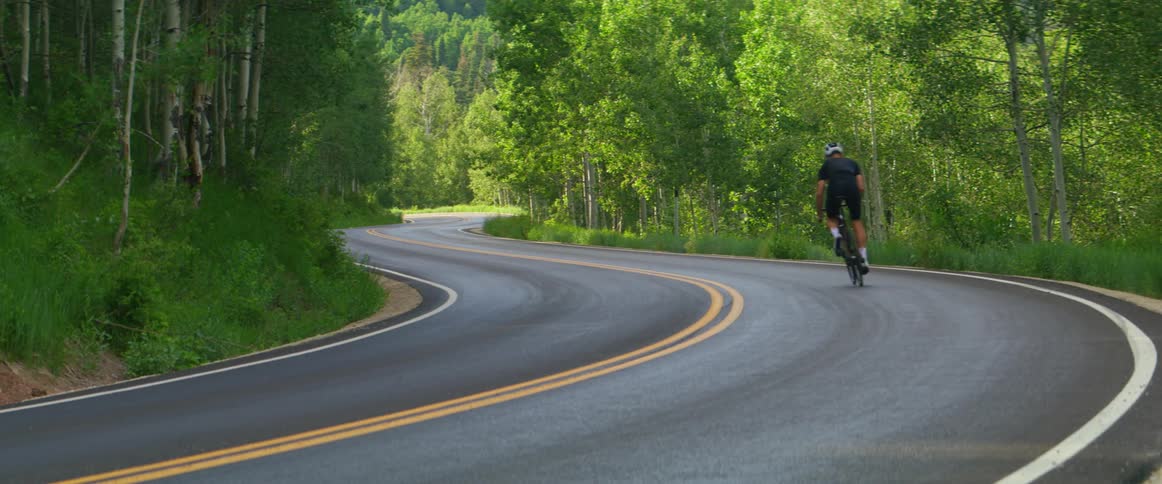 Man rides a bicycle around a bend in the highway next to a forest on a sunny day
