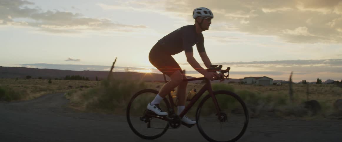 Man wearing a helmet and sunglasses rides a bicycle on a dirt road next to a field