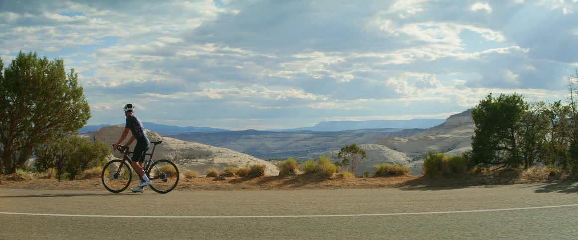 A man wearing a helmet is riding a bicycle on a road overlooking mountains as he stops and reaches for his water bottle