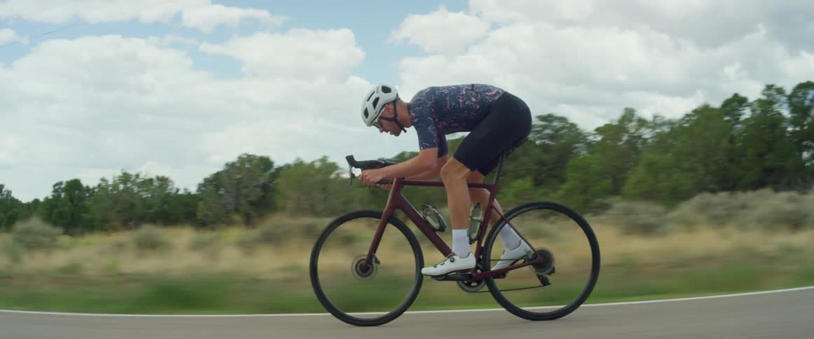A man wearing a white helmet is riding a bicycle on a road with a mountain in the distance and he leans over the handlebars as he gains speed