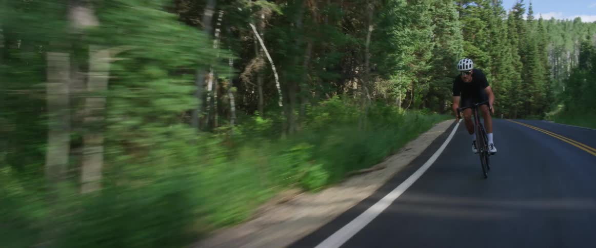 A man wearing a white helmet is riding a bicycle on a winding road through a forest
